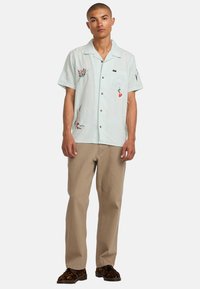 Chemise bleu clair à manches courtes avec broderies florales, boutons et poche poitrine, associée à un pantalon beige et des chaussures à imprimé animal.