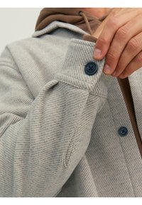 Szara marynarka w jodełkę z zapiętym mankietem na guzik i brązową bluzą z kapturem pod spodem. Teksturowana tkanina z subtelnymi detalami.