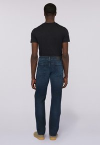 Hombre de espaldas con una camiseta negra, jeans azules con bolsillos traseros y zapatos beige, de pie contra un fondo claro y liso.