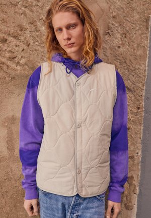 Beige kviltet vest med knaplukninger, båret over en lilla tie-dye hættetrøje. Har et let, struktureret stof.