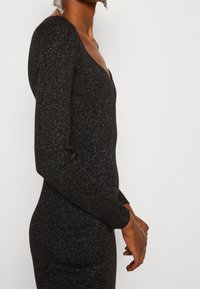 Abito lungo nero a maniche lunghe con scollatura profonda a girocollo, caratterizzato da un tessuto testurizzato e glitterato e una silhouette aderente. Dettagli includono maniche a costine.