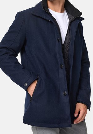 Man wearing a navy blue buttoned and zipped coat with hand in zippered pocket over a white shirt and grey pants.