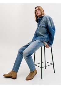 Άνδρας που φοράει μπλε τζιν μπουφάν και τζιν παντελόνια με καφέ σουέντ μπότες, κάθεται σε ψηλό μαύρο μεταλλικό σκαμπό μπροστά σε απλό φόντο.