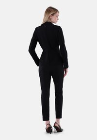 Schwarzes maßgeschneidertes Blazer mit passender Silhouette, lange Ärmel, strukturierter Stoff, mit Knopfdetails, kombiniert mit schlanken schwarzen Hosen und Absatzschuhen.