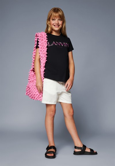 T-shirt noir avec un graphique "Lanvin Paris", shorts blancs, sac à épaule texturé rose et sandales noires sur une personne se tenant contre un fond gris.