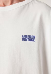 Valge puuvillane T-särk ümar kaelusega, vasakul rinnal sinise trükitud logoga "AMERICAN VINTAGE". Sujuv tekstuur.