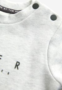 Szara bawełniana bluza z czarnymi guzikami na ramieniu i nadrukowanymi literami. Miękka tekstura, casualowy design, okrągły dekolt.