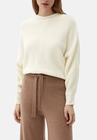 Cremefarvet ribstrikket sweater med rund hals, parret med brune strikkede bukser med snøre i taljen. Teksturerne er bløde og hyggelige.