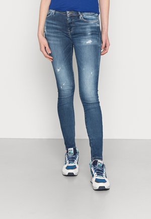 Jeans skinnys de ganga azuis com áreas desbotadas e pequenos detalhes de desgaste, combinados com ténis brancos e azuis com atacadores estampados.