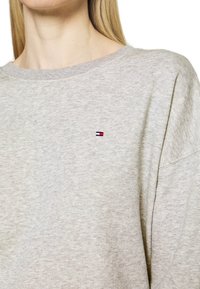 Primo piano di una persona che indossa un maglione grigio chiaro a collo rotondo con un piccolo logo rettangolare rosso, bianco e blu sul petto.