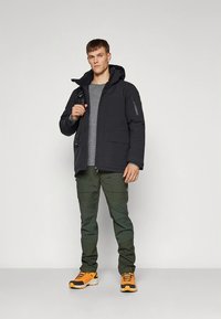 Juoda paminkštinta striukė su gaubtu, po ja pilka marškinėliai, žali kariniai kelnės ir oranžiniai sportbačiai. Striukėje yra užtrauktuku užsegama kišenė ant rankovės.