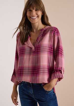 Femme souriante aux cheveux bruns portant une chemise à carreaux rose et violette avec les manches retroussées et un jean bleu, main dans la poche.