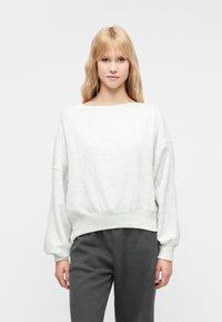 Svijetlo siva dukserica s širokim izrezom, spuštenim ramenima i rebrastim rubom. Materijal izgleda mekano i teksturirano. Uparena s tamno sivim hlačama.