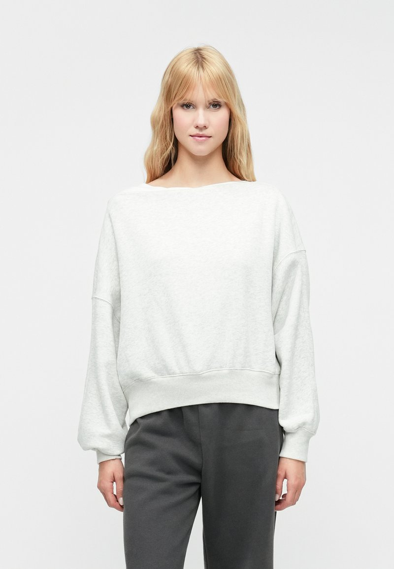 Svijetlo siva dukserica s širokim izrezom, spuštenim ramenima i rebrastim rubom. Materijal izgleda mekano i teksturirano. Uparena s tamno sivim hlačama.