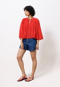 Rote, locker sitzende Bluse mit gerafftem Ausschnitt, kombiniert mit hoch taillierten Jeansshorts und flachen roten Sandalen. Lässiges Sommeroutfit.