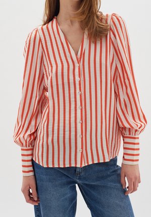 Kobieta ubrana w białą bluzkę z pionowymi czerwonymi paskami, zapinaną z przodu na guziki, z bufkami na długich rękawach, połączoną z niebieskimi dżinsami.