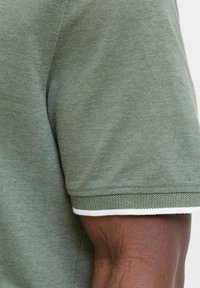 Zielona polo z fakturą, z białymi wykończeniami przy mankietach, prezentująca gładki, tkany materiał i klasyczny krój kołnierzyka.