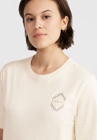 Camiseta de algodón color crema con mangas cortas, que presenta un logotipo gráfico en el lado izquierdo del pecho. Textura suave y cuello redondo clásico.