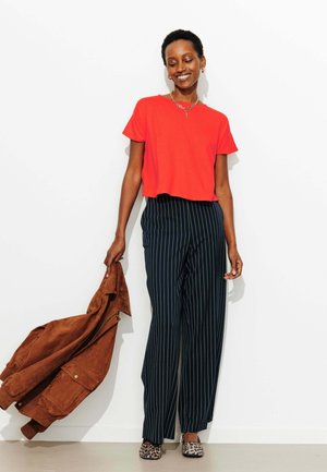 Femme souriante, portant un haut rouge à manches courtes, un pantalon noir à rayures fines, des chaussures imprimé léopard, tenant une veste marron dans sa main gauche.