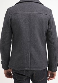 Hombre vestido con una chaqueta de lana gris oscuro y jeans vaqueros azul oscuro, visto desde atrás sobre un fondo liso.