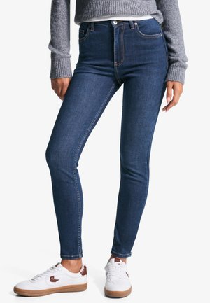 Dunkelblaue Skinny Jeans mit hoher Taille, die kontrastierende Nähte, einen vorderen Knopfverschluss und eine glatte Textur aufweisen. Tragen mit weißen Turnschuhen.
