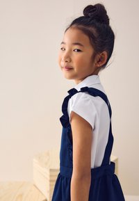 Jeune fille aux cheveux foncés attachés en chignon, portant une chemise blanche et une robe bleu marine, debout de profil sur un fond clair uni.