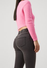 Person som bär en rosa fluffig kort tröja och åtsittande mörkgrå jeans, står på sidan mot en plain ljus bakgrund.