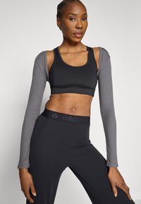 Svart sports-BH med grå lange ermer; glatt tekstur, tettsittende design. Svarte leggings med teksturert midjebånd med logo.