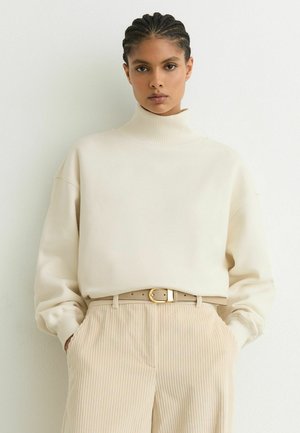 Vrouw met ingevlochten haar die een crèmekleurige coltrui draagt, ingestopt in een beige ribfluwelen broek met handen in de zakken, tegen een effen witte muur.