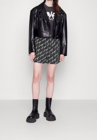 Veste en cuir noire courte, t-shirt graphique gris, mini-jupe verte à motifs et bottines noires épaisses à semelles robustes.
