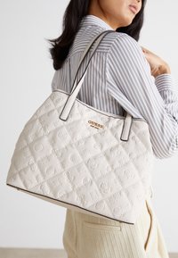 Biała pikowana torebka z wytłaczanymi wzorami logo, wyposażona w dwa długie uchwyty i kontrastujące czarne wykończenie. Złote logo z przodu.