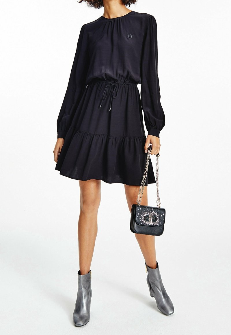 Schwarzes, knielanges Kleid mit langen Puffärmeln, gerafftem Ausschnitt, taillierter Taille und gerafftem Saum. Silberne Kettenhandtasche und graue Knöchelstiefel.