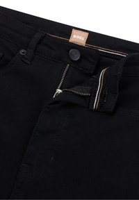 Pantalones vaqueros negros de mezclilla con cremallera y cierre de botón, y etiqueta con el logotipo. Presentan detalles de costura en blanco y marrón claro en la cinturilla.