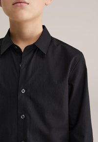 Melna pogu krekls ar plašu apkakli, garām piedurknēm un priekšējo joslu ar tumšām pogām; gluda tekstūra, minimālistisks dizains.