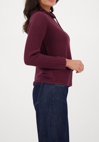 Bordeaux ribbed sweater met een capuchon, lange mouwen en zijsplitten, gecombineerd met donkerblauwe denim jeans. De stof heeft een gestructureerde uitstraling.