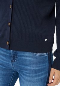 Marineblaue gerippte Strickjacke mit braunen Knöpfen, aus strukturiertem Stoff und kleinem Markenlogo am Saum, kombiniert mit blauen Jeans.