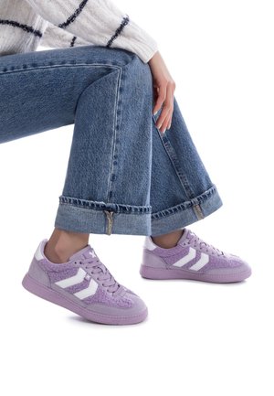 Zapatillas moradas hechas de ante y tejido texturizado, con franjas blancas, puntera redonda y suela de goma. Combinadas con unos vaqueros de pierna ancha.