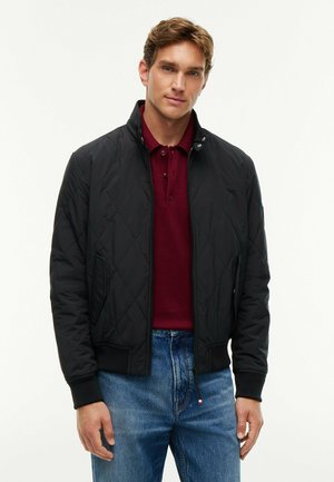 Mand iført en sort quiltet jakke over en bordeaux polo og blå jeans, stående mod en ensartet lys baggrund.