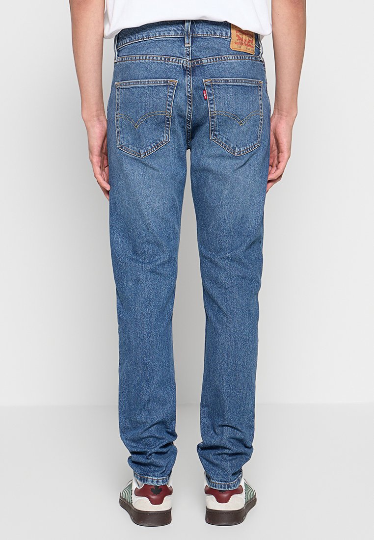 Blaue Denim-Jeans mit geradem Schnitt, ausgestattet mit einem braunen Lederetikett, zwei Gesäßtaschen und einer ausgewaschenen Textur. Kombiniert mit weißen und kastanienbraunen Sneakers.