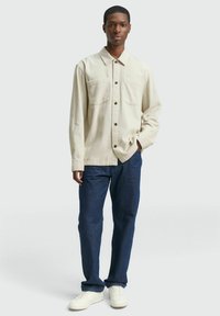 Lichtbeige katoenen overhemd met twee borstzakken, gecombineerd met donkerblauwe rechte spijkerbroek en witte sneakers. Stikdetails zichtbaar.