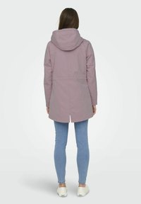 Hellrosa, wasserdichte Jacke mit Kapuze, mit tailliertem Schnitt, langen Ärmeln und einem längeren Rückensaum, kombiniert mit hellblauen Jeans und weißen Schuhen.