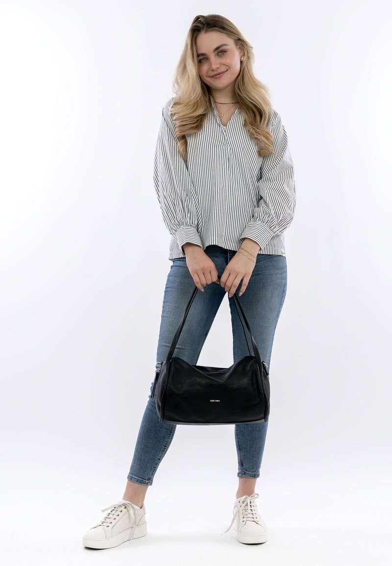 Schwarze Lederhandtasche mit weicher, runder Form und einer dezent strukturierten Oberfläche. Das Modell trägt ein gestreiftes Oberteil und blaue Jeans.