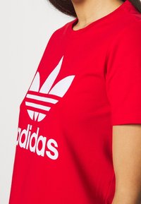 Červené bavlněné tričko s výrazným bílým logem Adidas a designem se třemi pruhy na přední straně, s klasickým kulatým výstřihem a krátkými rukávy.