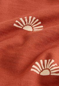 Terrakotta-stof med et solstrålemønster i off-white, der kendetegnes ved en blød tekstur med let glans og tydelig vævedetalje.