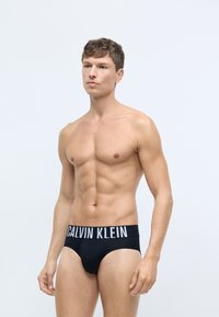 Čierne bavlnené slipsy so širokým bielym pásom s logom "CALVIN KLEIN". Majú priliehavý strih a hladkú textúru, ktorá zdôrazňuje postavu.