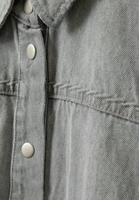 Grå denimskjorte med sølvfarvede trykknapper, spids krave og synlige syningsdetaljer langs fronten og skuldrene.