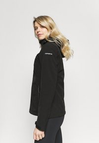 Zwarte softshell jas met een slim fit ontwerp, zijzakken en een capuchon. Het materiaal lijkt glad met een subtiele glans en heeft logo-accenten.