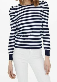 Maglia a righe blu navy e bianca con maniche a sbuffo, scollatura rotonda e polsini a coste, abbinata a jeans bianchi. Tessuto morbido, design casual.