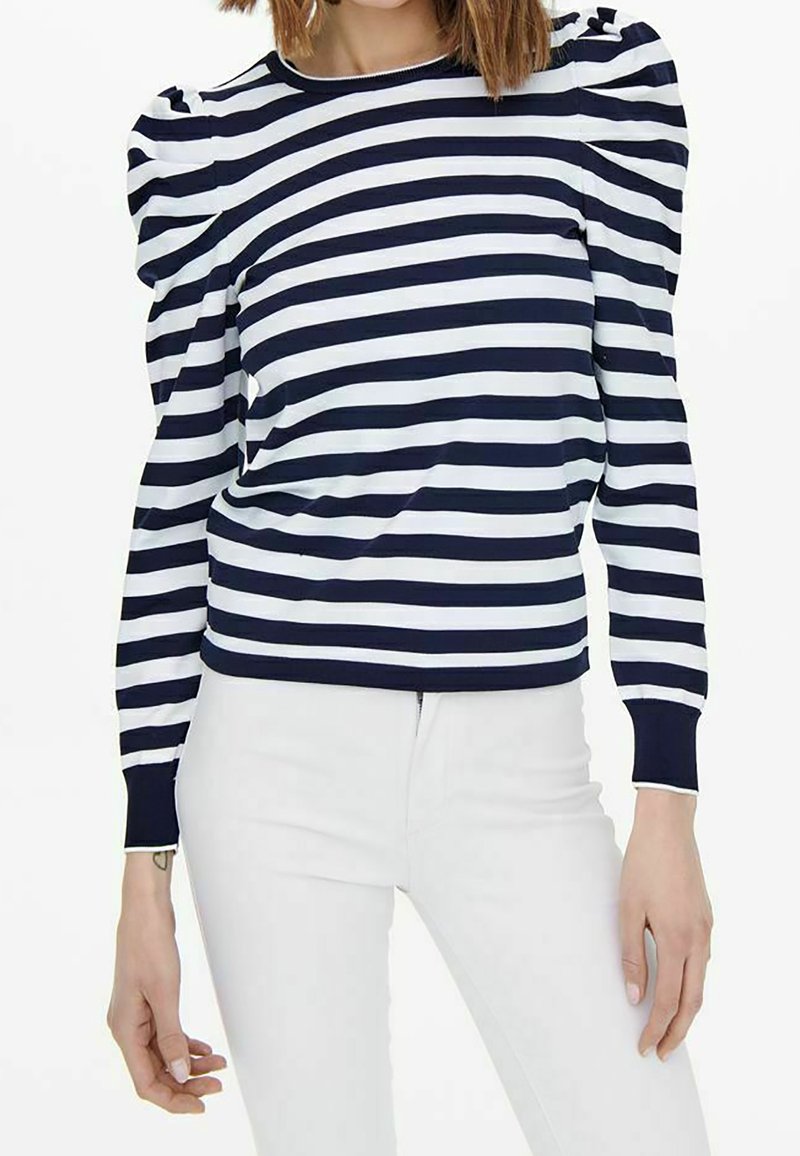 Maglia a righe blu navy e bianca con maniche a sbuffo, scollatura rotonda e polsini a coste, abbinata a jeans bianchi. Tessuto morbido, design casual.