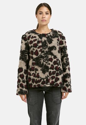 Woman wearing buttoned, textured beige jacket with black and dark red abstract floral patterns, paired with faded black jeans.